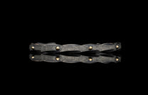 Dark hammered metal bracelet with golden rivets and a twisted design, reflected on a black surface.