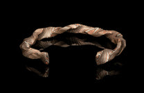 Wavy metal cuff bracelet with coppery and silver striped patterns, on a reflective black surface.