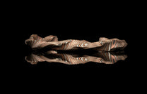 Wavy layered jewelry band with bronze and silver stripes, reflected on a black surface.