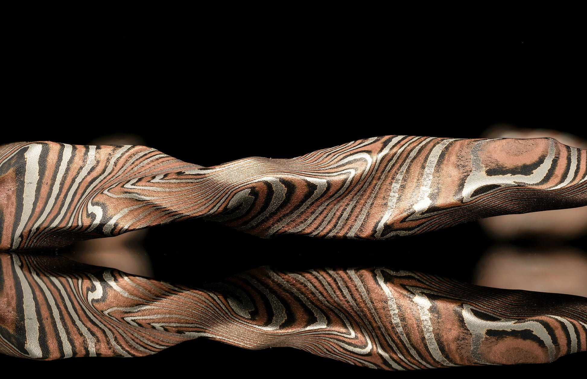 Sculptural jewelry piece with wavy organic stripes in copper, silver, and dark metal, reflected on black.
