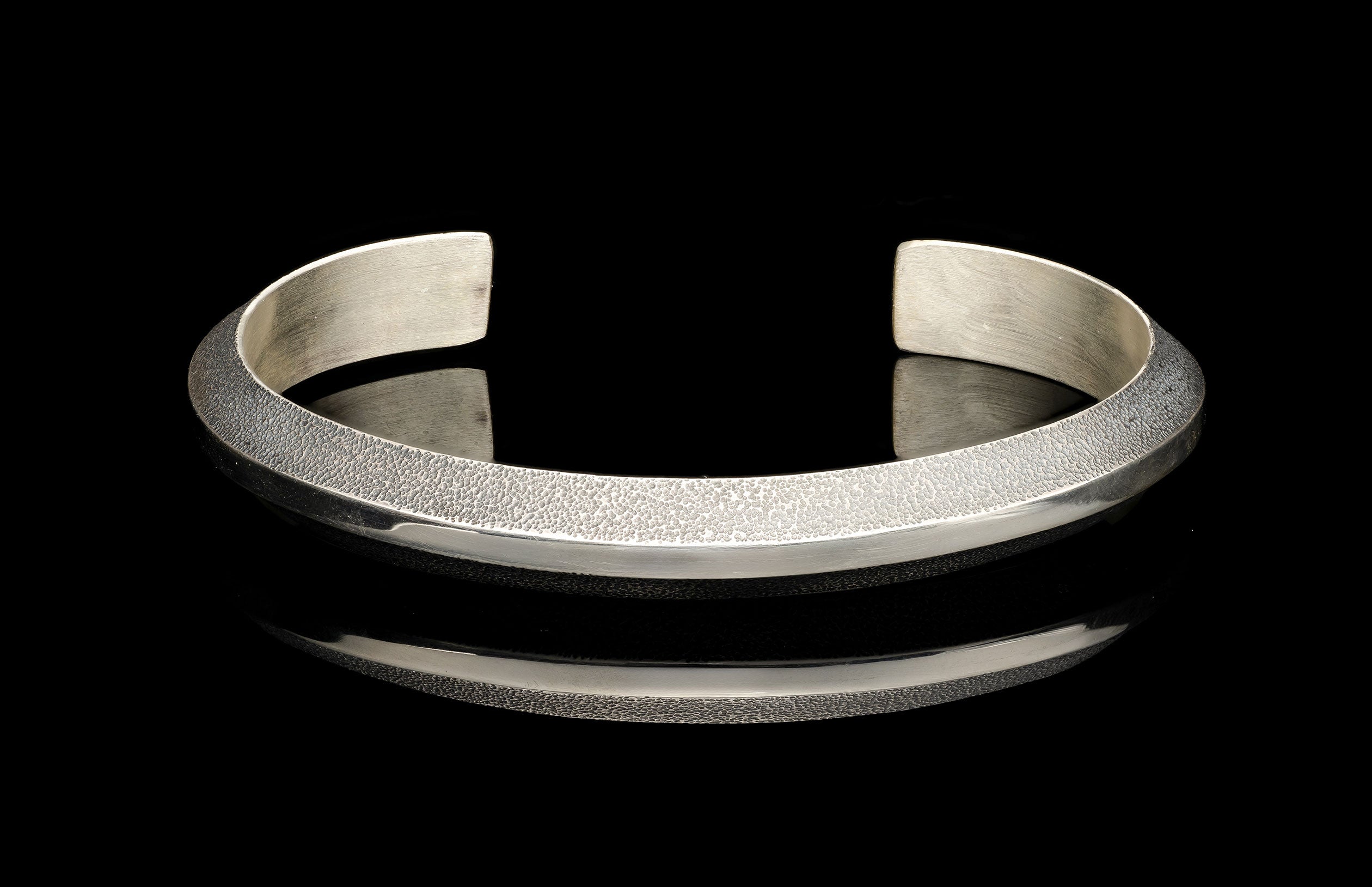 Silver cuff bracelet with a hammered texture on its upper band and a polished lower band, on a reflective black surface.
