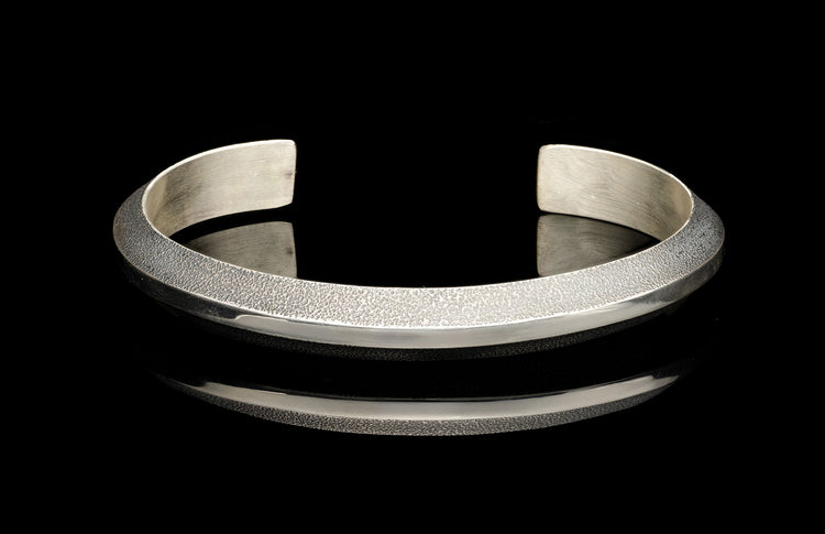 Silver cuff bracelet with a hammered texture on its upper band and a polished lower band, on a reflective black surface.