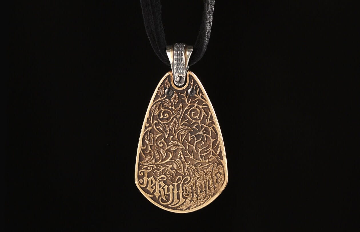 Bronze teardrop pendant with intricate embossed leaves, vines, and "Jekyll + Hyde" text, on a black cord.