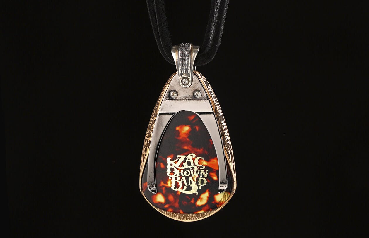 Bronze and silver-toned pendant with engraved edges and 'William Henry' inscription, holding a tortoiseshell guitar pick with a gold Zac Brown Band logo, on a black cord.