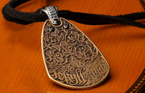 Bronze 'Jekyll + Hyde' pendant featuring ornate vine engraving and black stones, on a black cord, resting on wood.