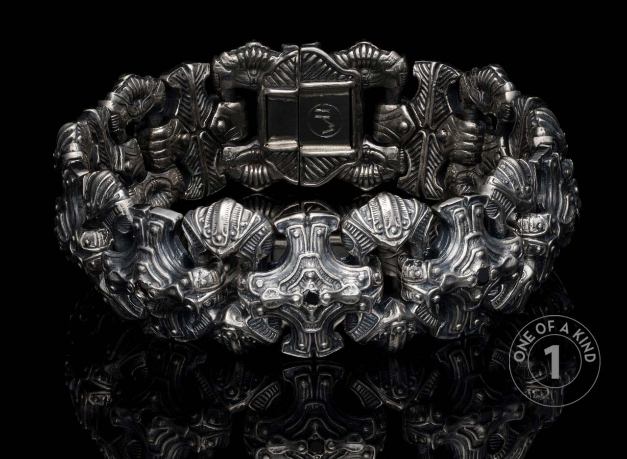 Silver-toned metal bracelet with intricate, ornate skull-like and armored links, detailed engravings, on a reflective black surface.
