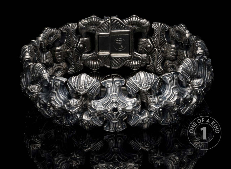 Silver-toned metal bracelet with intricate, ornate skull-like and armored links, detailed engravings, on a reflective black surface.