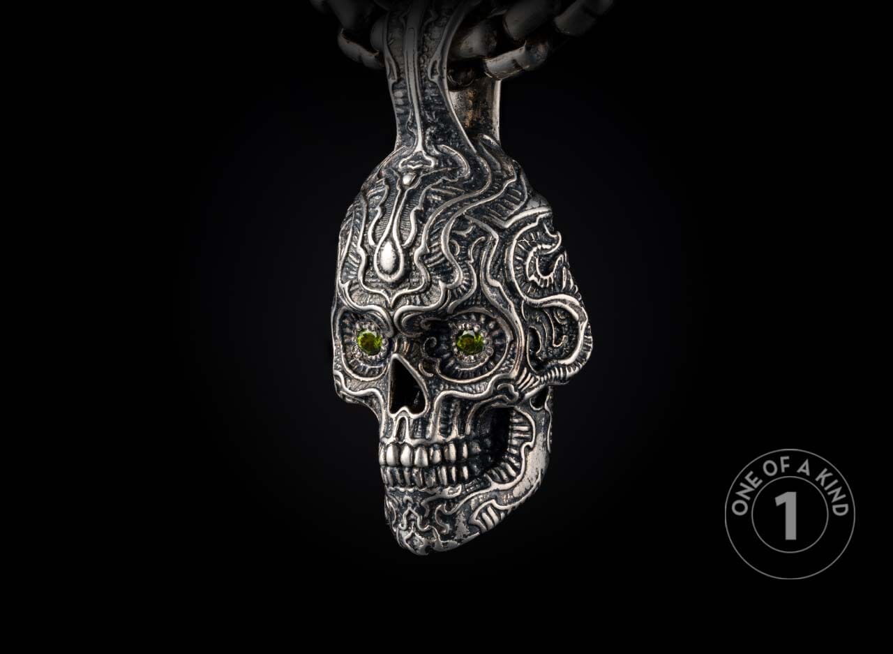 Ornate silver skull pendant with intricate tribal carvings, green gemstone eyes, hanging from a metal chain.