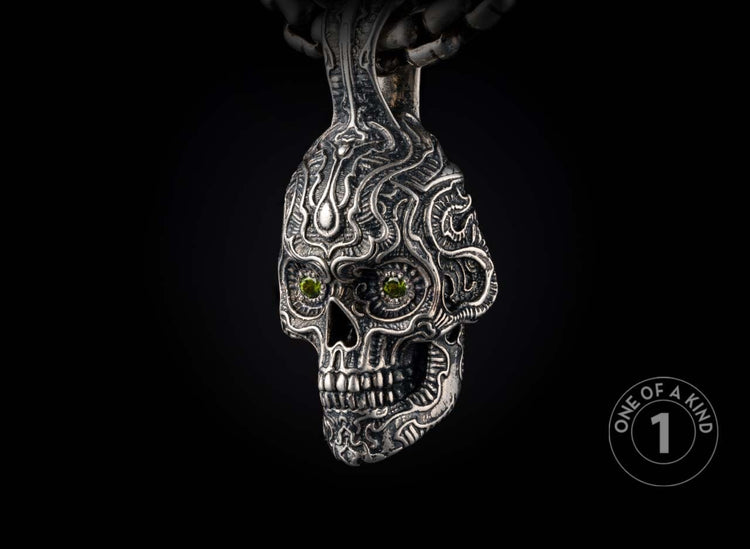 Ornate silver skull pendant with intricate tribal carvings, green gemstone eyes, hanging from a metal chain.