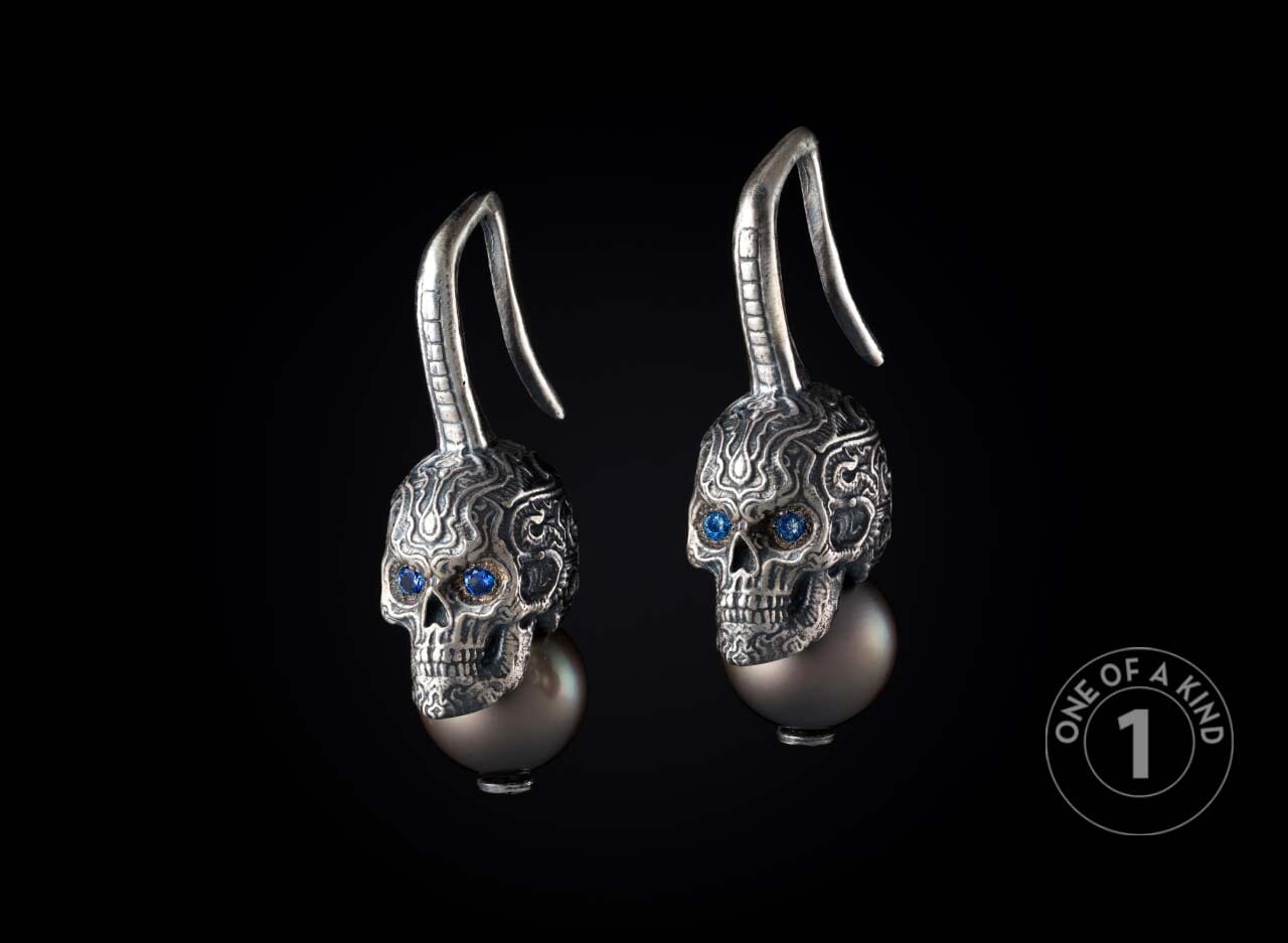 Pair of silver skull earrings with intricate carved patterns, blue gem eyes, and dark grey pearl drops.