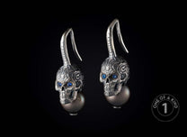 Pair of silver skull earrings with intricate carved patterns, blue gem eyes, and dark grey pearl drops.