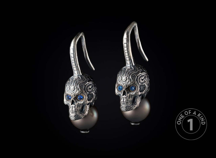 Pair of silver skull earrings with intricate carved patterns, blue gem eyes, and dark grey pearl drops.