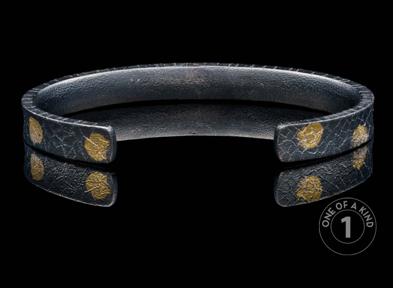 Dark textured metal cuff bracelet with irregular gold leaf-like spots, reflected on a black surface.