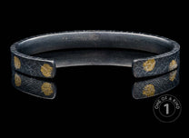 Dark textured metal cuff bracelet with irregular gold leaf-like spots, reflected on a black surface.