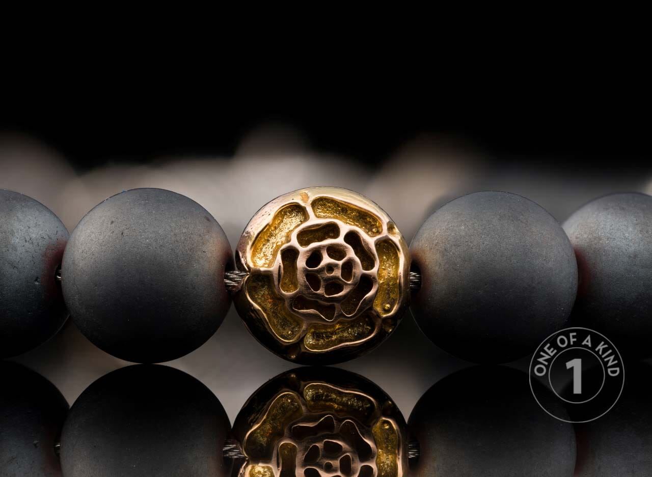 Matte dark grey beaded bracelet with a central gold-toned metal bead featuring a textured swirling design, reflected below.