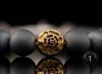 Matte dark grey beaded bracelet with a central gold-toned metal bead featuring a textured swirling design, reflected below.