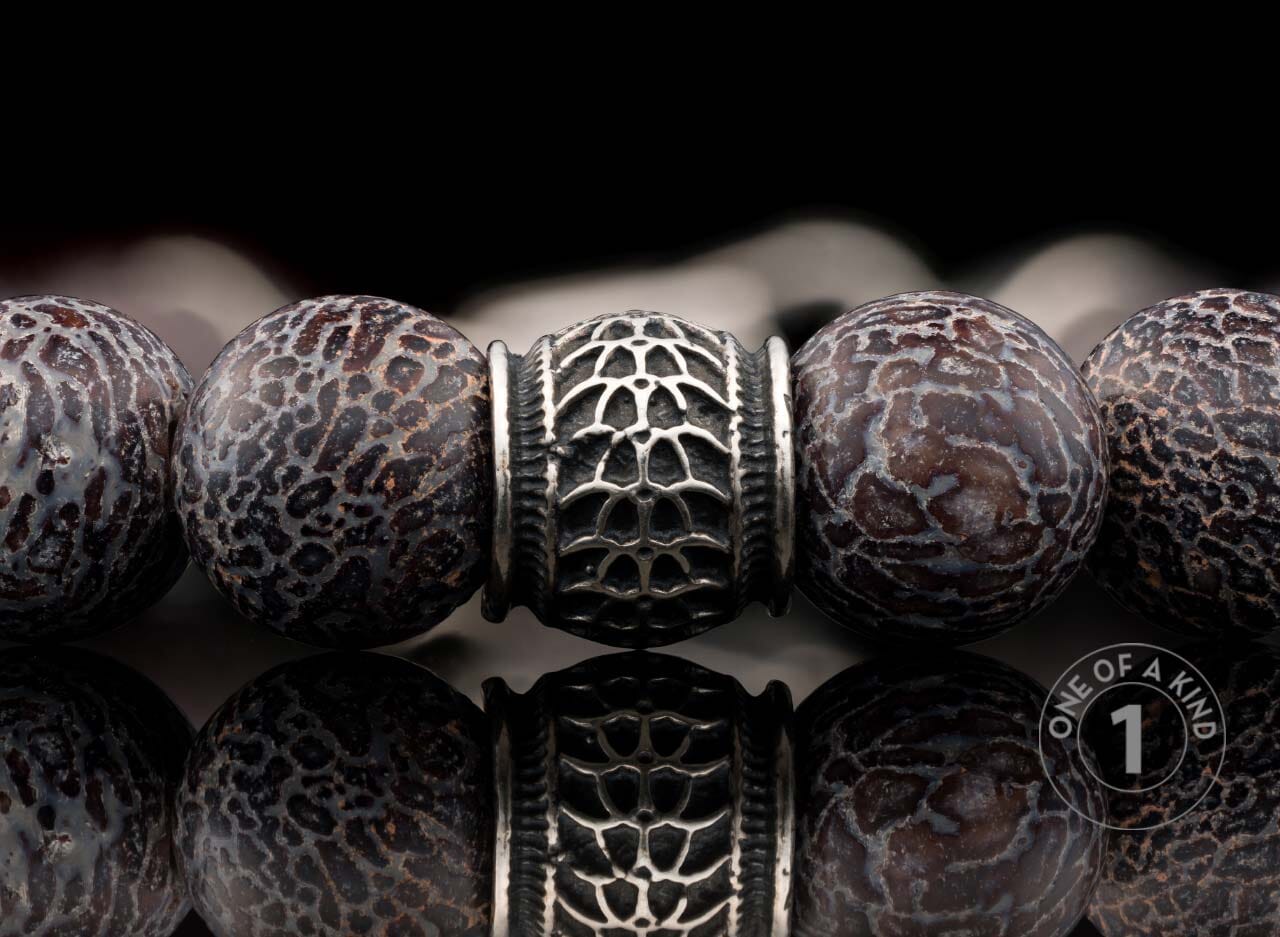 Beaded bracelet with dark brown patterned stone beads and a silver ornate metal bead, reflected below.