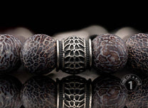 Beaded bracelet with dark brown patterned stone beads and a silver ornate metal bead, reflected below.