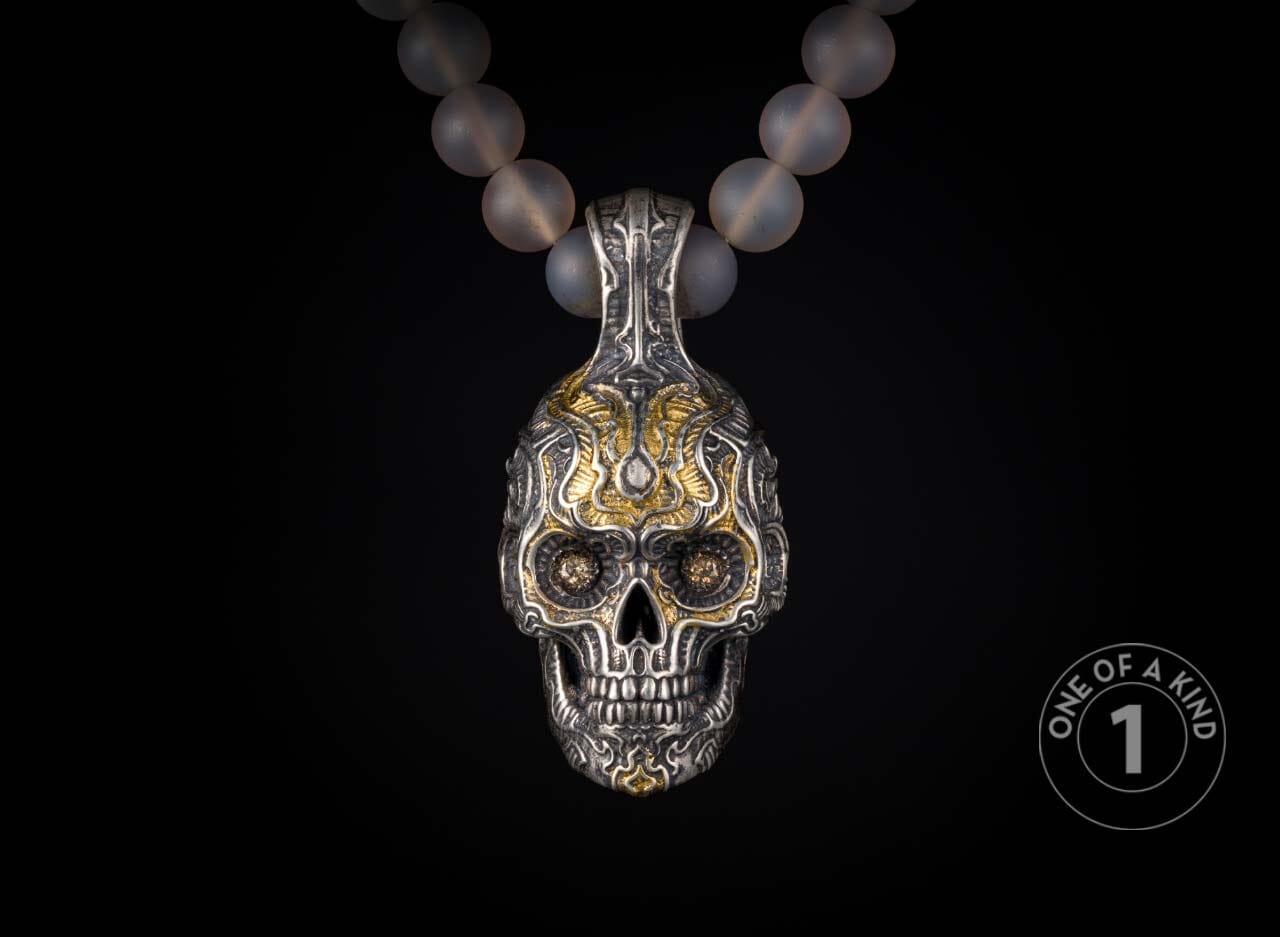Ornate silver and gold skull pendant with intricate carvings and dark gemstone eyes, suspended from a translucent beaded necklace.