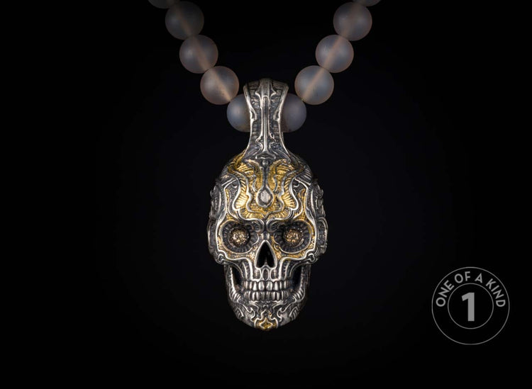 Ornate silver and gold skull pendant with intricate carvings and dark gemstone eyes, suspended from a translucent beaded necklace.