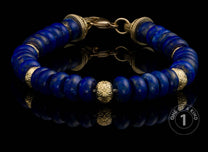 Dark blue lapis lazuli beaded bracelet with gold-tone carved spacers and clasp, resting on a reflective surface.