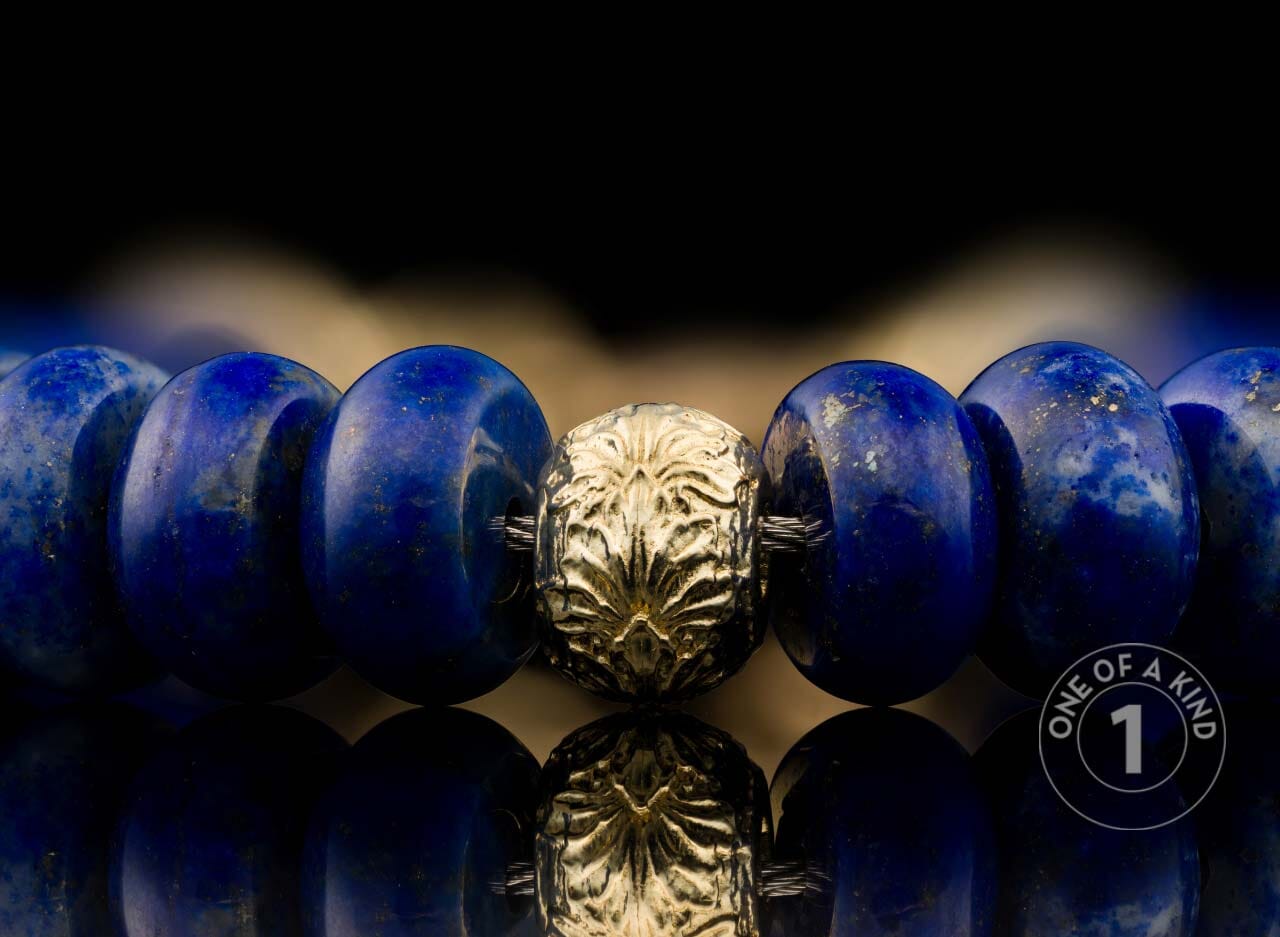 Beaded bracelet with deep blue lapis lazuli beads and a central textured gold-tone bead, reflected on a dark surface.
