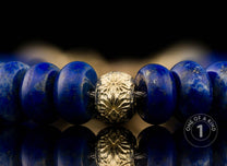 Beaded bracelet with deep blue lapis lazuli beads and a central textured gold-tone bead, reflected on a dark surface.