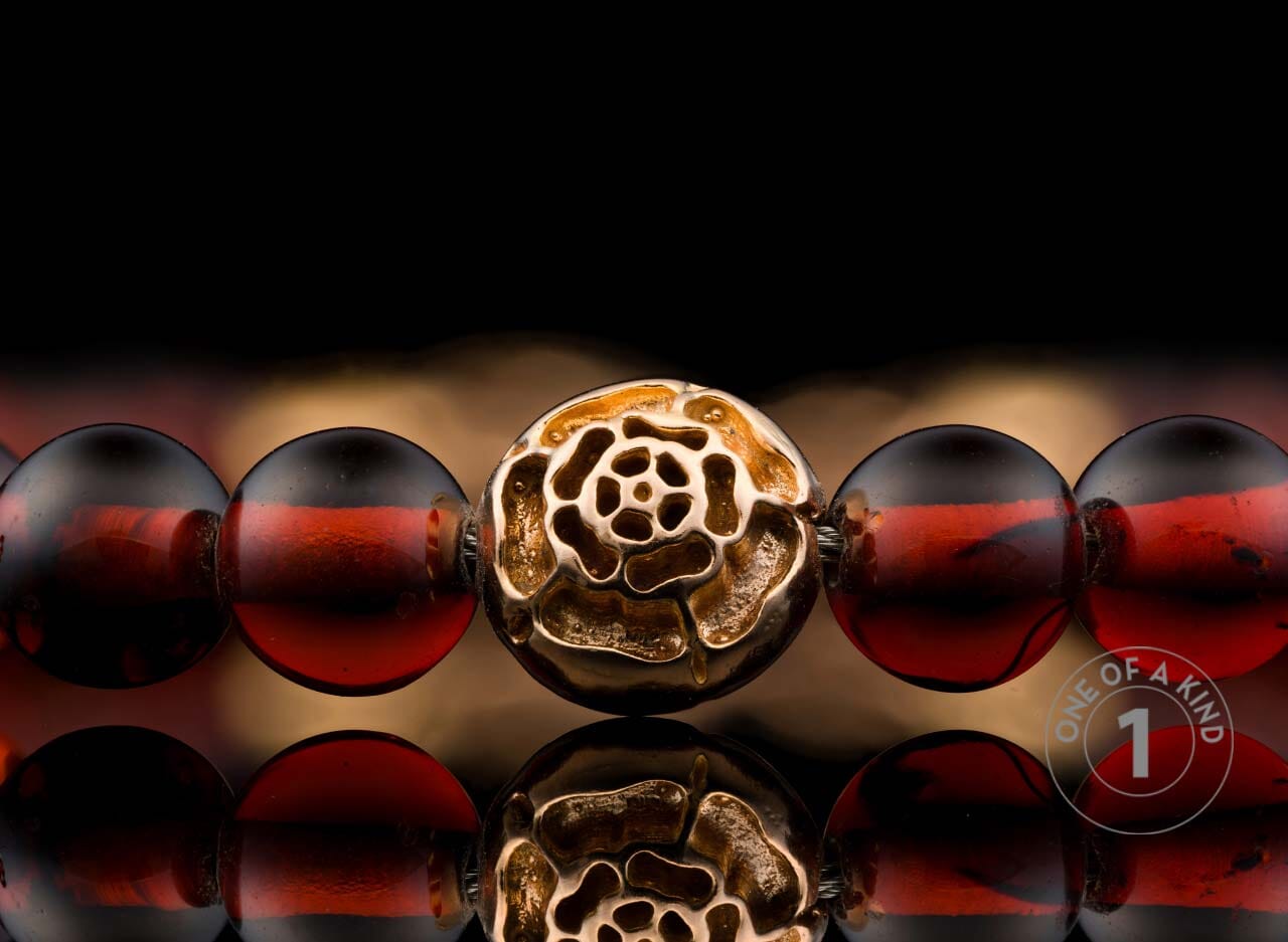 Beaded bracelet with dark red beads and a detailed golden rose-shaped centerpiece, shown close-up on a reflective surface.