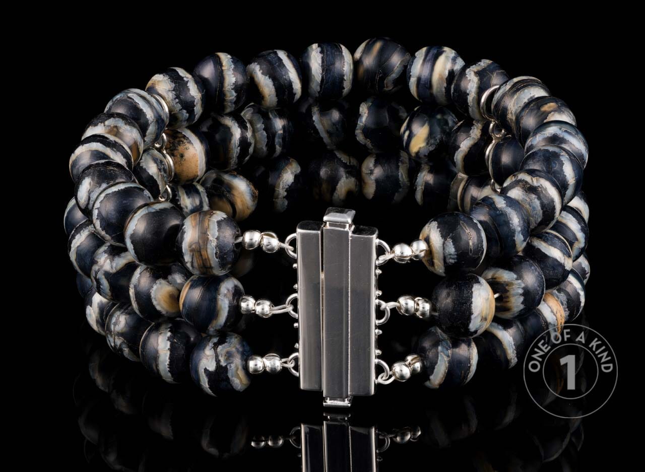 Three-strand bracelet with black, white, and tan banded agate beads and a silver-toned clasp, on a reflective black surface.