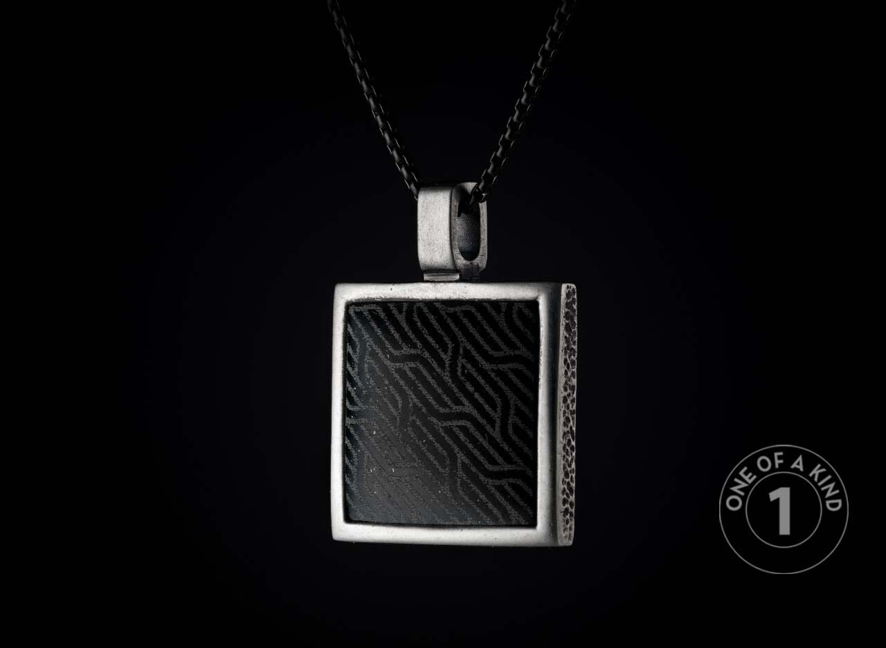 Square silver-toned metal pendant with a black insert featuring a wavy geometric pattern and textured sides, hanging from a dark chain.
