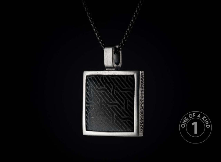 Square silver-toned metal pendant with a black insert featuring a wavy geometric pattern and textured sides, hanging from a dark chain.