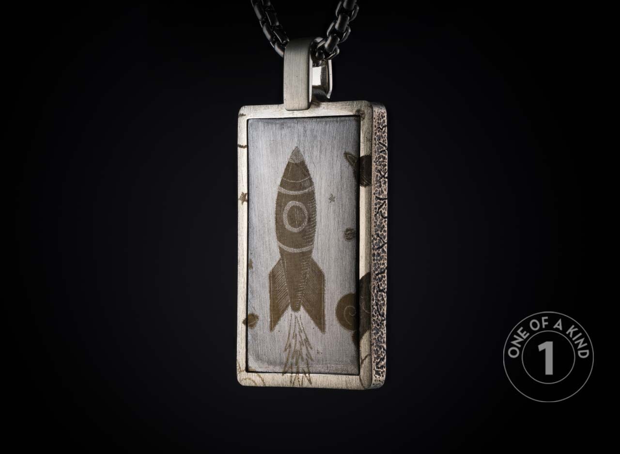 Oxidized silver rectangular pendant featuring an engraved rocket, stars, and textured frame on a dark chain.