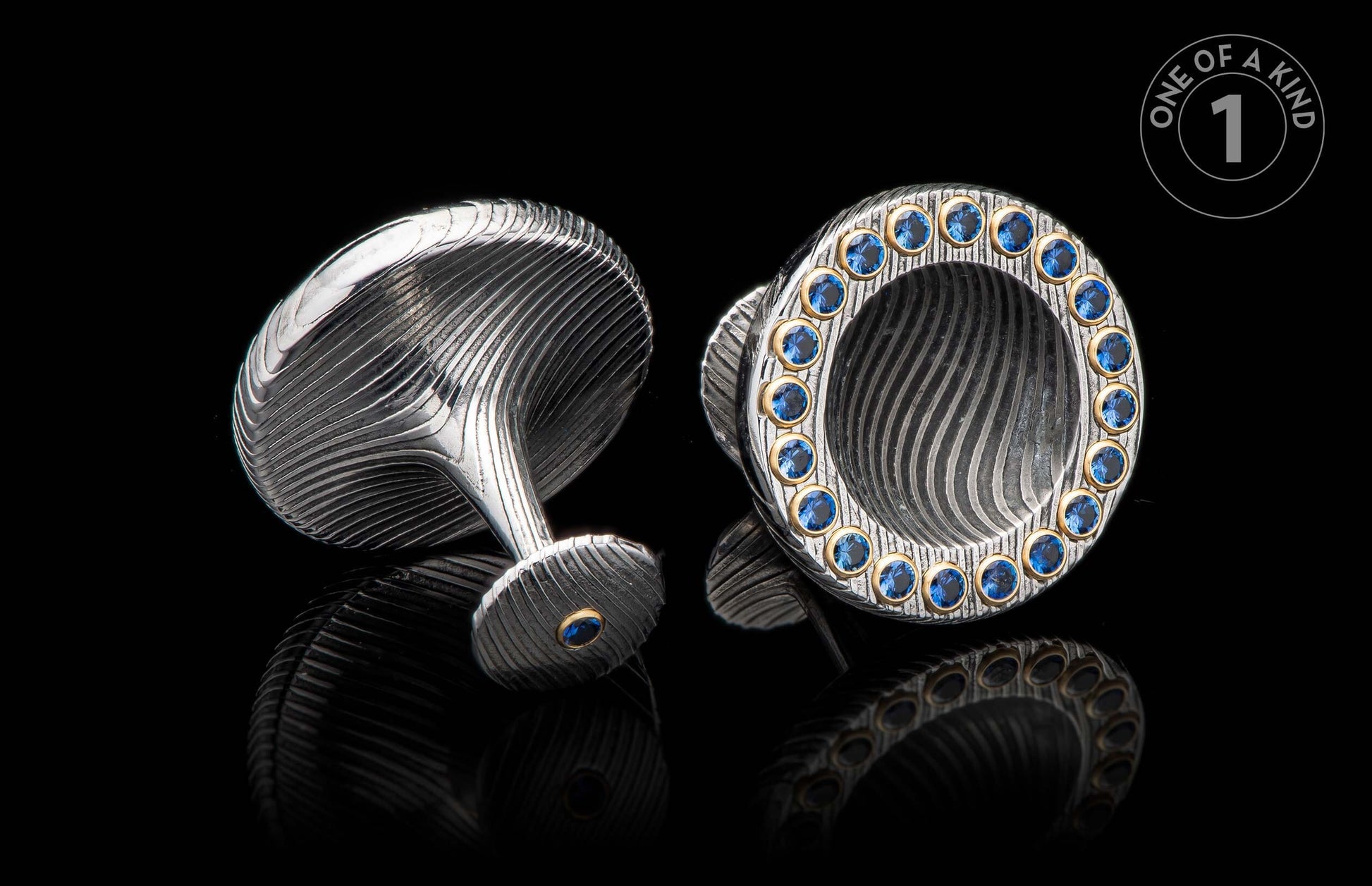 Silver cufflinks with wavy texture, one featuring multiple blue gemstones, the other with a single blue gem, all in gold settings, on a reflective black surface.