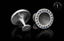 Silver cufflinks with wavy texture, one featuring multiple blue gemstones, the other with a single blue gem, all in gold settings, on a reflective black surface.