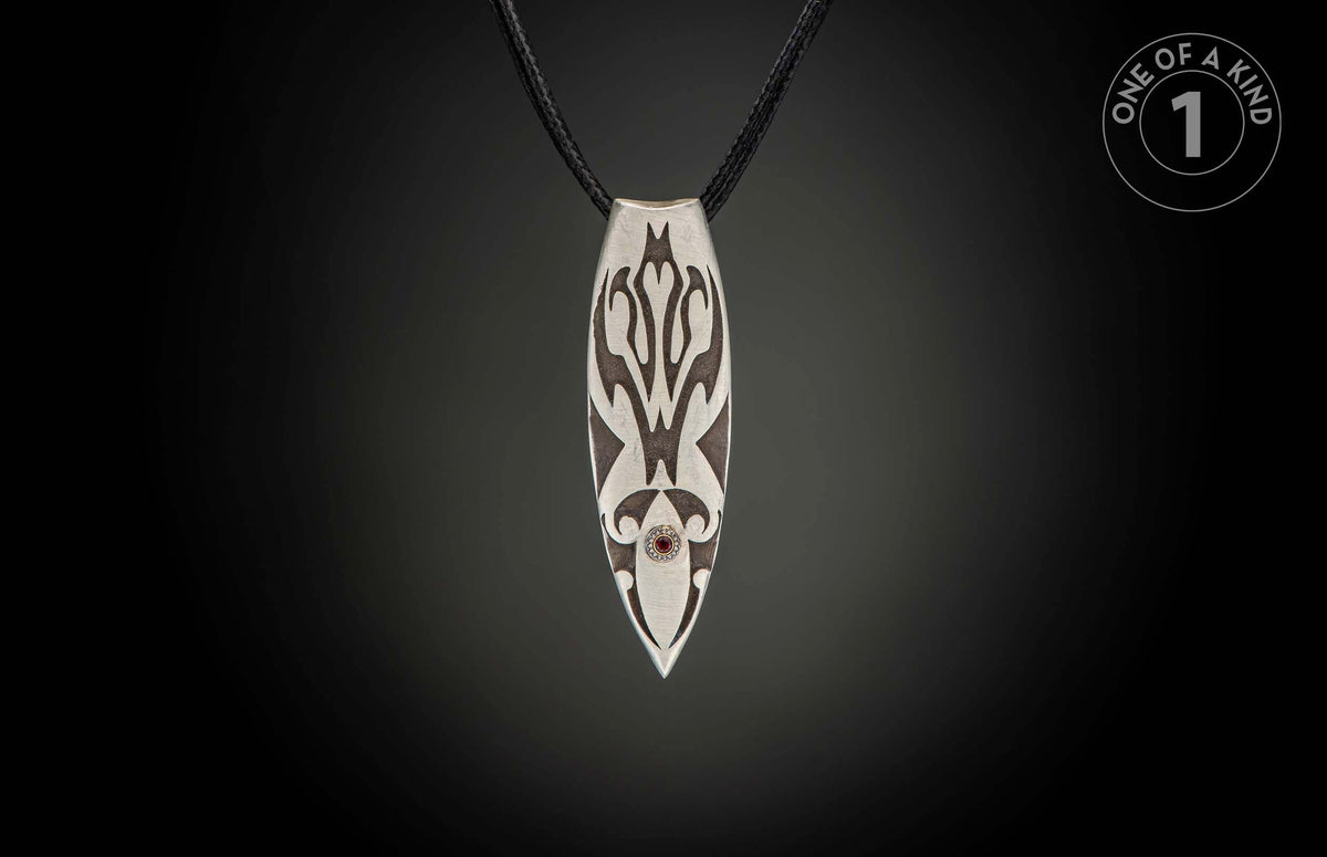 Silver pendant with dark etched patterns, a red gemstone, and black braided cord.