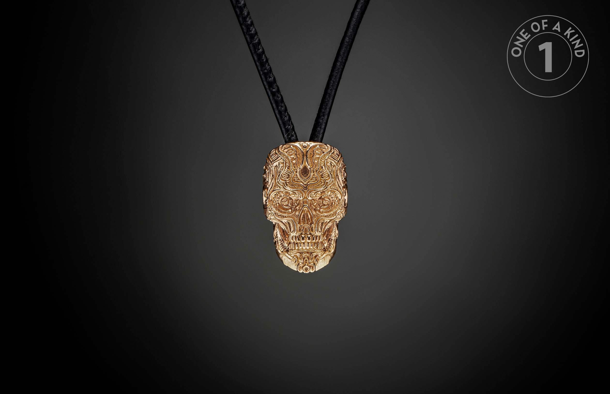 Golden skull pendant with intricate relief patterns, hanging from black cords, and a 'ONE OF A KIND' label.