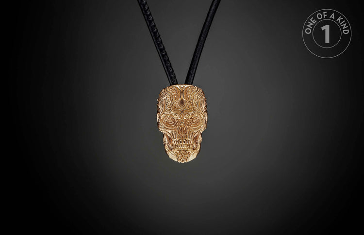 Golden skull pendant with intricate relief patterns, hanging from black cords, and a 'ONE OF A KIND' label.