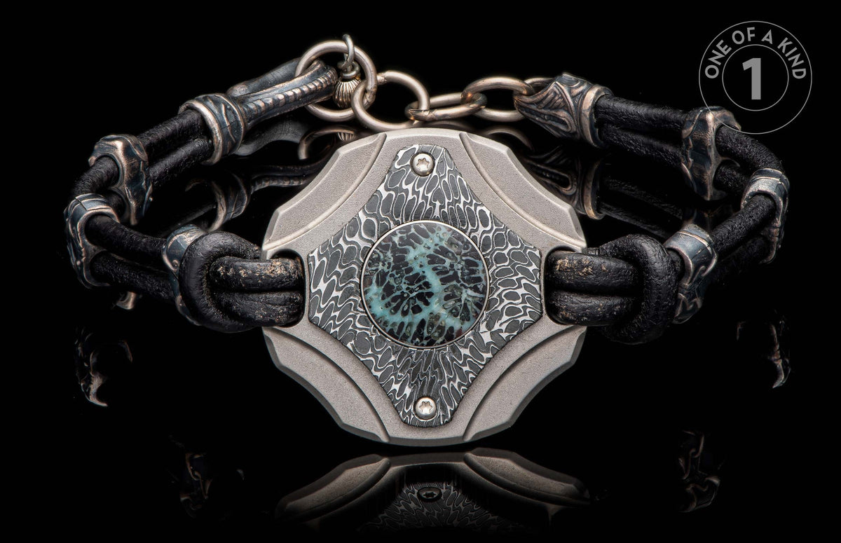 Black leather bracelet with textured metal links and an intricate patterned centerpiece with a blue-green round inlay.