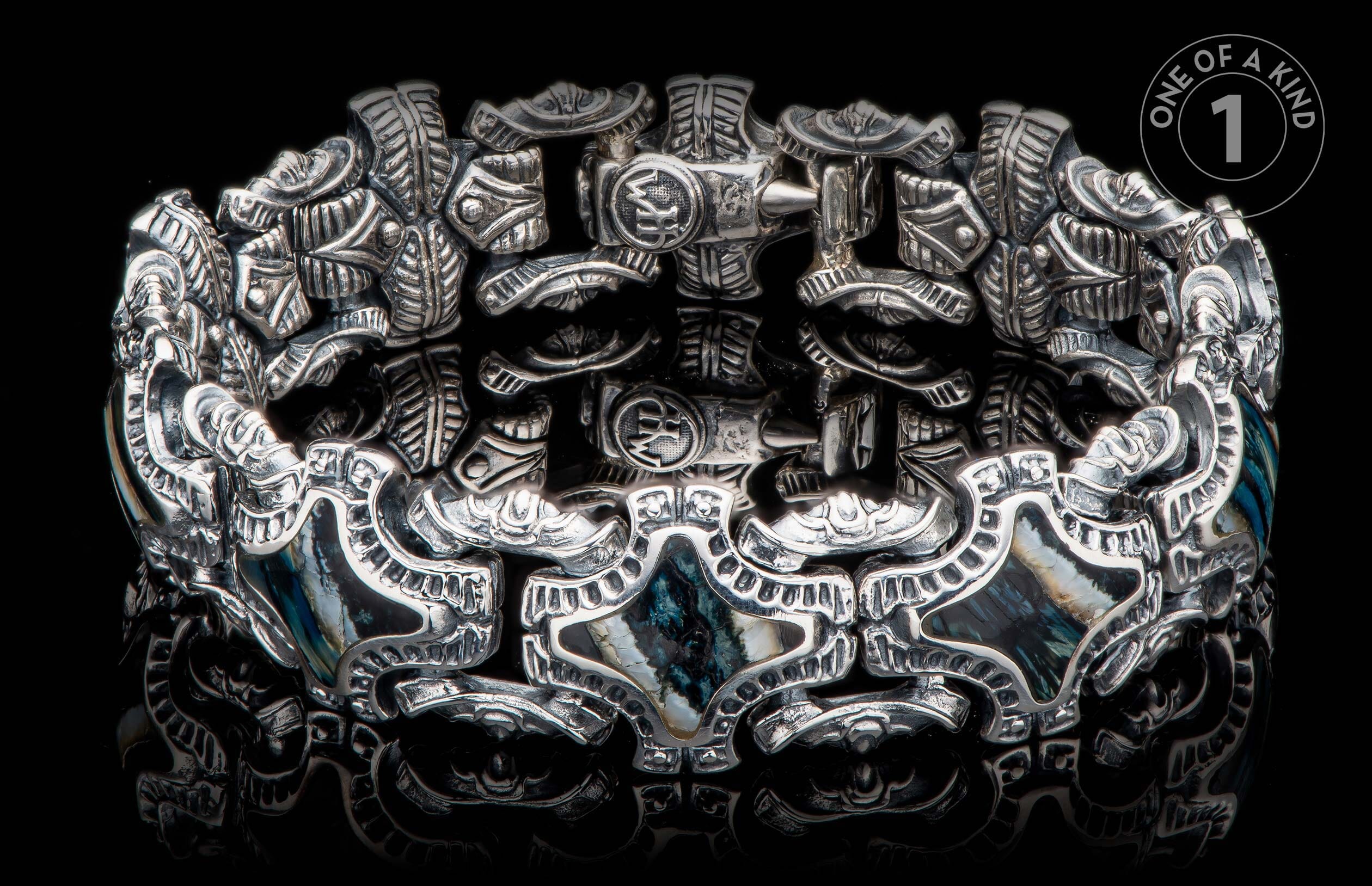 Silver bracelet with intricate carved patterns and dark blue-green shell inlays on a reflective black surface.