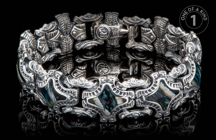 Silver bracelet with intricate carved patterns and dark blue-green shell inlays on a reflective black surface.