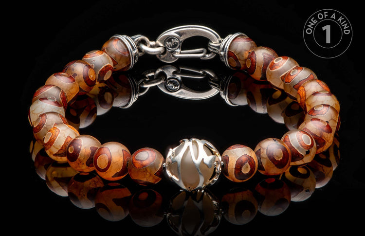 Beaded bracelet with brown and cream eye-patterned stones, a silver-toned clasp, and an ornate silver centerpiece on a reflective black surface.