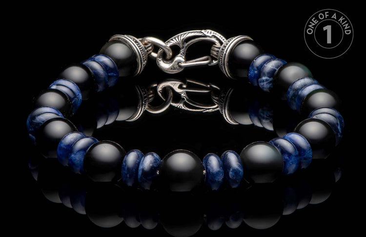 Black matte and blue patterned bead bracelet with an ornate silver clasp on a reflective surface.