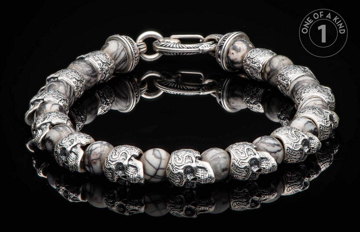 Silver-tone and marbled stone bracelet with skull beads and an ornate clasp, lying on a reflective black surface.