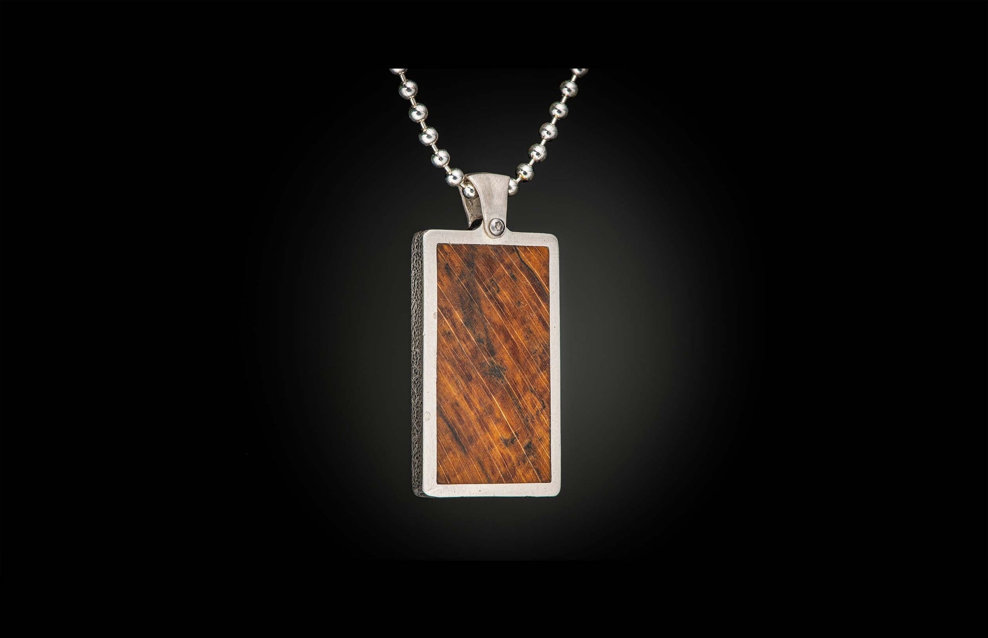 Rectangular pendant with a reddish-brown wood inlay featuring diagonal grain, set in a textured silver-tone metal frame, suspended from a ball chain.