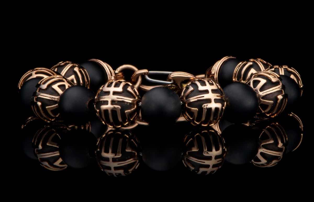 Black beaded bracelet with ornate gold-tone patterned beads and a metallic clasp, reflected on a dark surface.