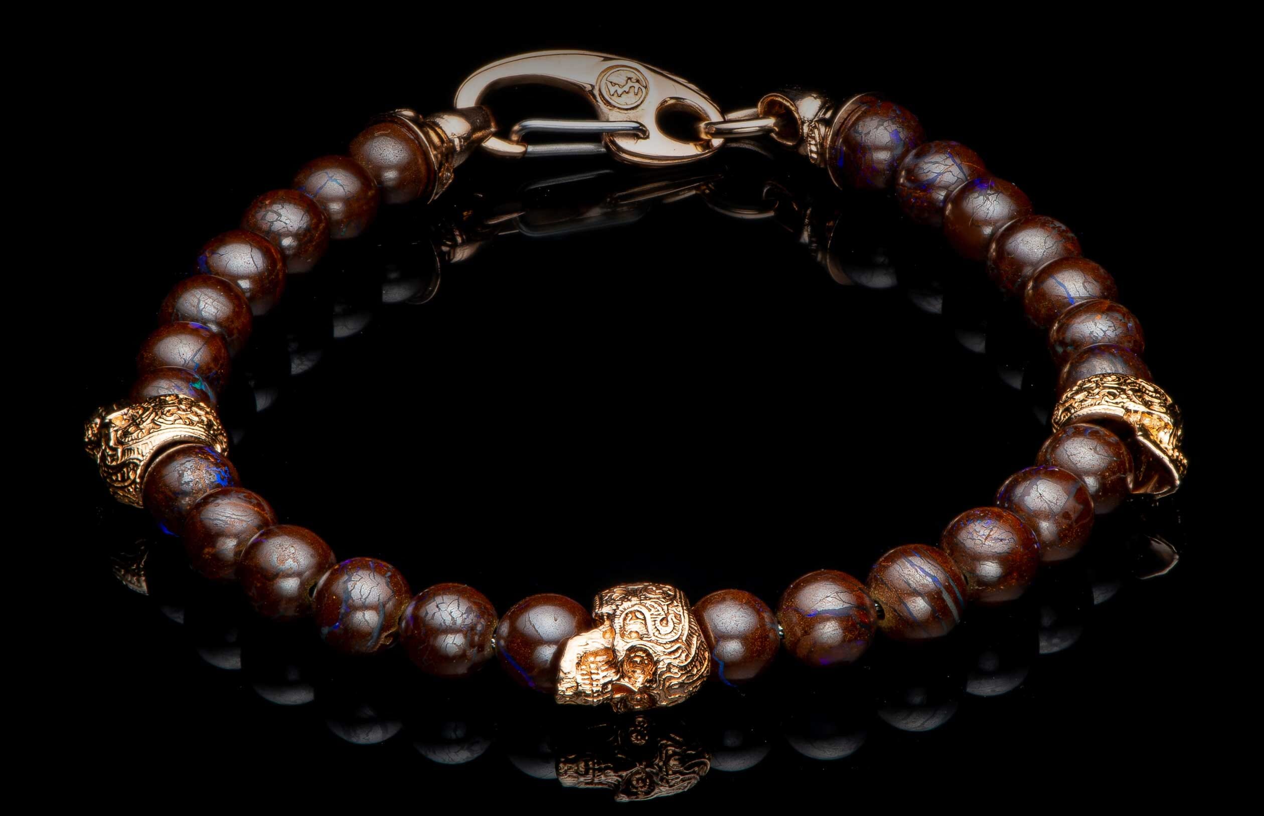 Brown beaded bracelet with blue flecks, gold-toned skull charms, and a hook clasp, reflecting on black.