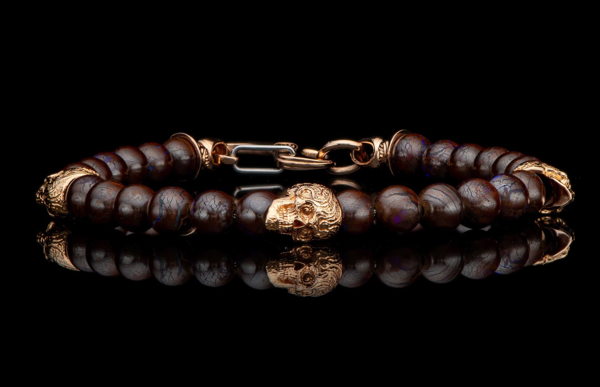 Dark brown beaded bracelet with a gold-toned skull bead and ornate clasp, reflecting on a black surface.