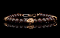Dark brown beaded bracelet with a gold-toned skull bead and ornate clasp, reflecting on a black surface.