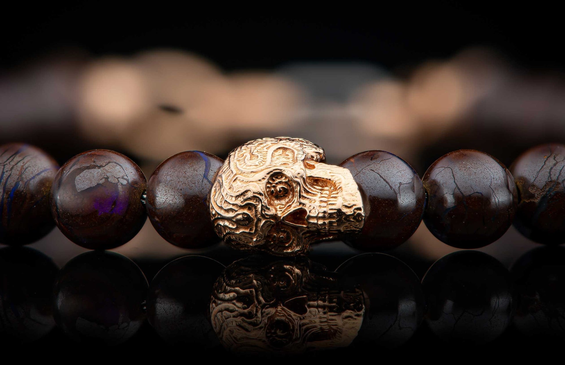 Dark brown beaded bracelet with a gold skull bead featuring ornate patterns and subtle blue veins, reflected on a dark surface.