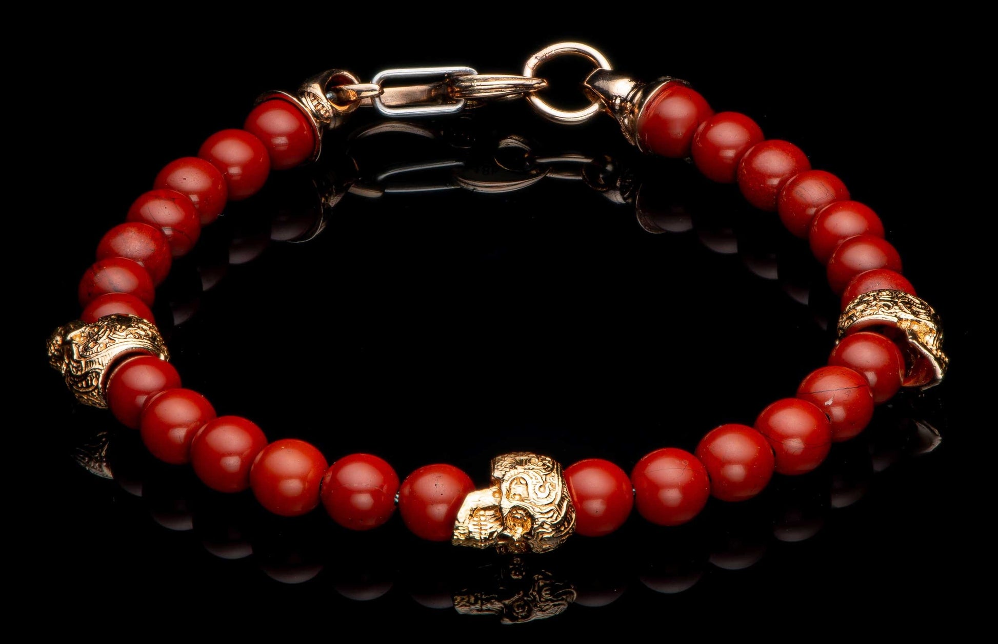 Red beaded bracelet with gold-toned skull beads and a gold and silver clasp on a black reflective surface.